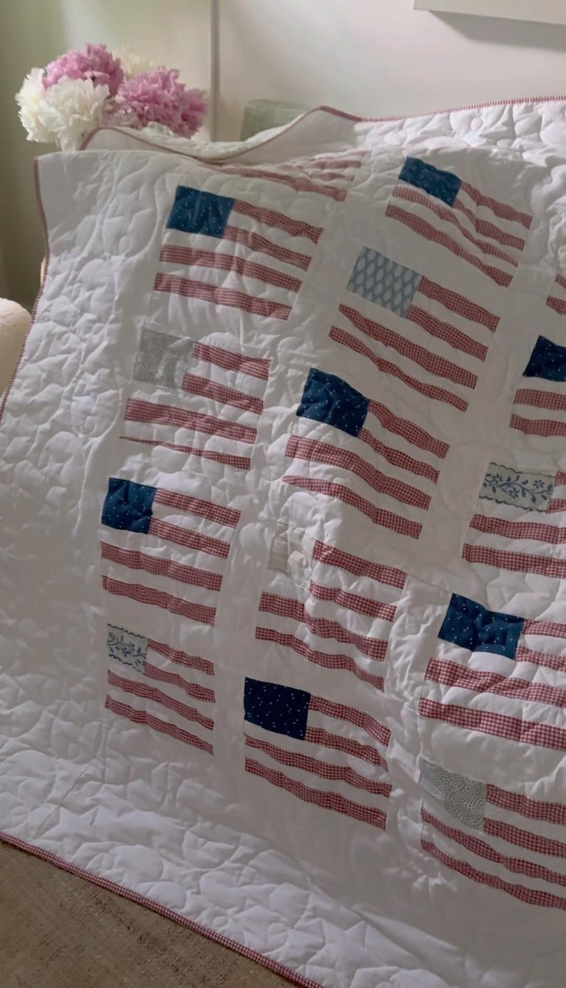 K&ouml;nnte beinhalten: Wei&szlig; gesteppte Decke mit mehreren amerikanischen Flaggenmotiven. Die Flaggen haben rot-wei&szlig; gestreifte Muster mit blauen quadratischen Feldern. Die Decke hat einen rot-wei&szlig; karierten Rand und ein Steppmuster.