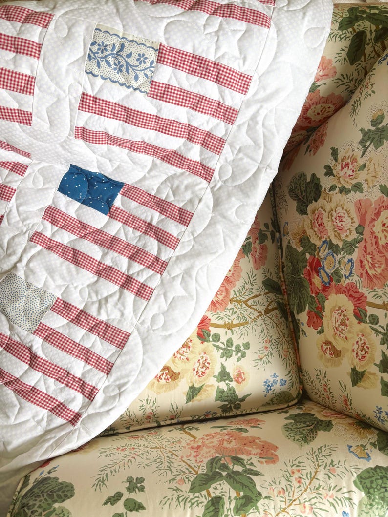 May include: A quilted blanket with a patriotic American flag design. The flag features red and white striped sections and blue sections with white stars or floral patterns. The blanket is on a floral patterned couch.
