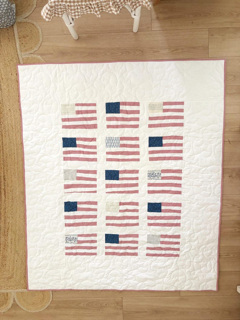 K&ouml;nnte beinhalten: Eine wei&szlig;e Steppdecke mit rotem Vichy-Rand und einem Muster aus amerikanischen Flaggen. Jede Flagge hat rot-wei&szlig;e Streifen und ein blaues Quadrat mit wei&szlig;en Sternen oder anderen Mustern. Die Steppdecke ist mit einem wellenf&ouml;rmigen Muster gesteppt.