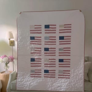 K&ouml;nnte beinhalten: Eine wei&szlig;e Steppdecke mit einem Muster aus amerikanischen Flaggen. Jede Flagge hat rot-wei&szlig;e Streifen und ein blaues Feld mit wei&szlig;en Sternen. Die Steppdecke ist mit einem rosa Rand versehen und &uuml;ber einem Sofa drapiert.
