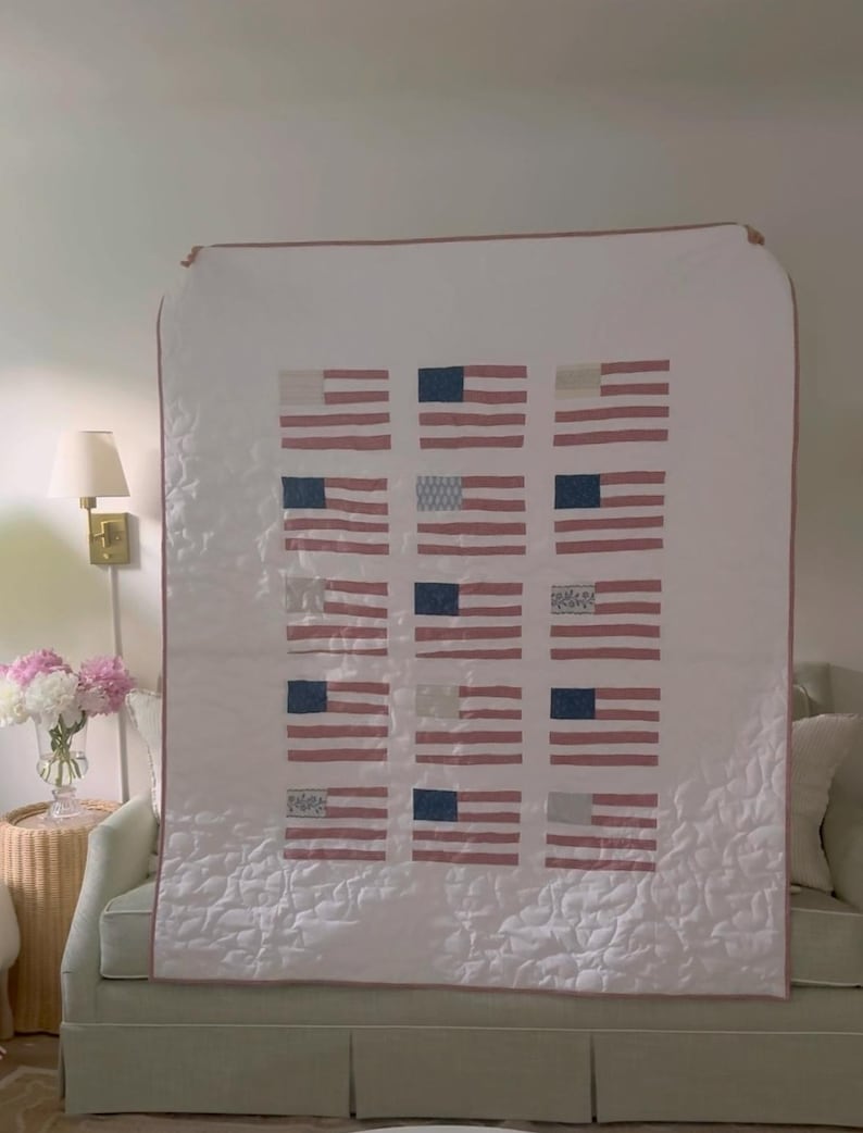 K&ouml;nnte beinhalten: Eine wei&szlig;e Steppdecke mit einem Muster aus amerikanischen Flaggen. Jede Flagge hat rot-wei&szlig;e Streifen und ein blaues Feld mit wei&szlig;en Sternen. Die Steppdecke ist mit einem rosa Rand versehen und &uuml;ber einem Sofa drapiert.