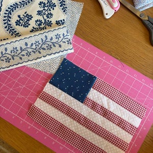 K&ouml;nnte beinhalten: Ein teilweise fertiggestellter amerikanischer Stoffflagge auf einer rosa Schneidematte. Die Flagge hat ein blaues Quadrat mit wei&szlig;en Sternen, rot-wei&szlig; gestreifte Abschnitte. Stoffreste, ein Rollschneider und eine Schere sind ebenfalls sichtbar.