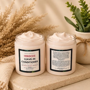 Puede incluir: Dos frascos de acondicionador sin enjuague de hibisco con crema blanca. Las etiquetas dicen "CONFESSIONSBYNAVEI" y "HIBISCUS LEAVE-IN CONDITIONER". Los frascos están sobre una superficie de piedra, con plantas secas y una pequeña planta en maceta en el fondo. Cada frasco contiene 227g.
