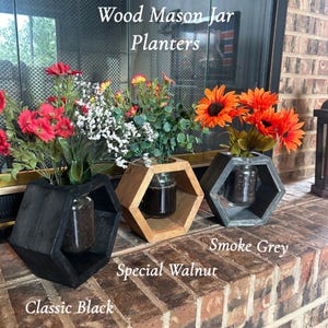 Pode incluir: Três floreiras hexagonais de madeira para potes de vidro, rotuladas como Classic Black, Special Walnut e Smoke Grey. Cada floreira contém um pote de vidro com flores artificiais, incluindo flores vermelhas, brancas e laranjas.