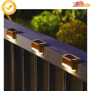 May include: Three solar-powered lights are mounted on a metal railing. The lights are rectangular with a bronze finish and emit a warm, inviting glow. The background is blurred, suggesting an outdoor setting.