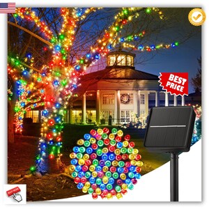 May include: A tree illuminated with colorful LED string lights, with a solar panel and a cluster of lights in the foreground. The lights are red, green, blue, and yellow. A building is in the background.