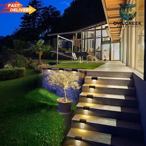 May include: Outdoor scene at dusk featuring a modern home with solar lights illuminating the steps and retaining wall. The lights are rectangular and emit a warm glow. The scene includes a lush green lawn, a small tree in a pot, and the text "OWL CREEK SOLAR LIGHTS".