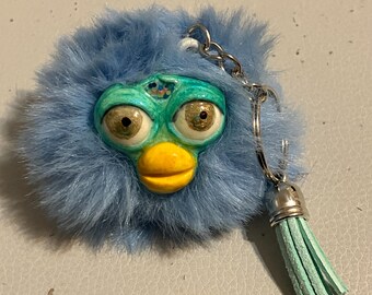 Könnte beinhalten: Ein blauer, flauschiger Schlüsselanhänger mit einem stilisierten Gesicht mit großen Augen, einem gelben Schnabel und einem türkisfarbenen Gesicht. Der Schlüsselanhänger hat eine silberne Kette und eine türkisfarbene Quaste.