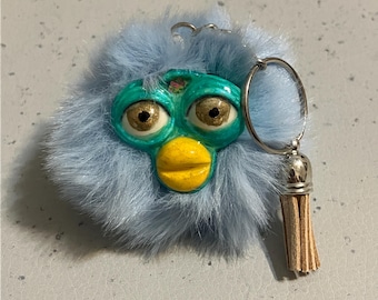 Könnte beinhalten: Ein von Furby inspirierter Schlüsselanhänger in Blau und Türkis mit flauschiger Oberfläche. Der Schlüsselanhänger hat einen silbernen Schlüsselring und eine kleine Quaste. Das Furby hat goldene Augen und einen gelben Schnabel.
