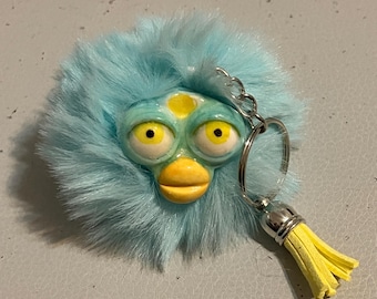 Könnte beinhalten: Ein skurriler Schlüsselanhänger mit einem von Furby inspirierten Gesicht, großen gelben Augen und einem gelben Schnabel. Das Gesicht ist von hellblauem Kunstfell umgeben. Ein silberner Schlüsselring und eine gelbe Quaste vervollständigen das Design.