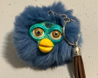 Könnte beinhalten: Ein blauer Fell-Schlüsselanhänger mit einem Furby-ähnlichen Gesicht, einem türkisfarbenen Gesicht, einem gelben Schnabel und braunen Augen. Der Schlüsselanhänger hat einen silbernen Ring und eine braune Quaste.