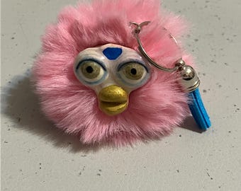 Könnte beinhalten: Ein pinkfarbener, flauschiger Schlüsselanhänger mit einem stilisierten Gesicht, großen Augen, einem goldenen Schnabel und einem blauen Akzent auf der Stirn. Der Schlüsselanhänger hat einen silbernen Reifen und eine kleine blaue Quaste.
