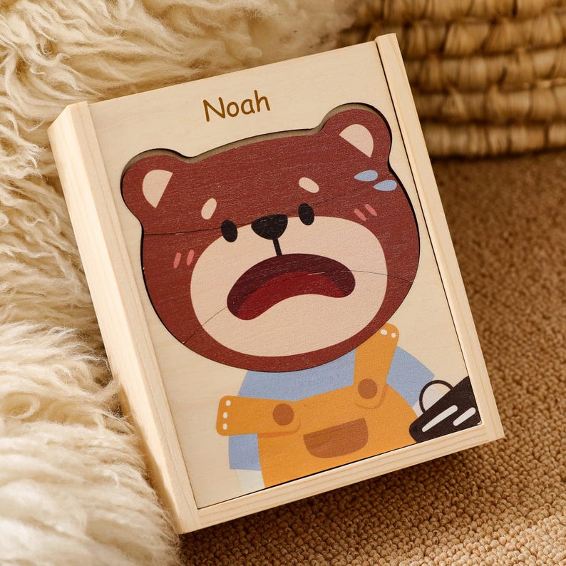 K&ouml;nnte beinhalten: Holzpuzzle-Box mit einem betr&uuml;bten B&auml;rengesicht und dem Namen "Noah" oben. Der B&auml;r hat einen braunen Kopf, rosa Wangen und ein blaues Hemd mit gelben Latzhosen. Das Puzzle befindet sich in einer hellen Holzkiste.