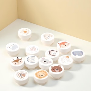 May include: A selection of white cabinet knobs, each adorned with charming illustrations of animals and nature elements. Designs include a sun, rainbow, fox, deer, koala, bear, lion, and elephant. These knobs are ideal for adding a playful touch to any furniture.