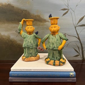 May include: Two ceramic monkey figurines, each wearing a green robe and a yellow hat. One monkey holds a dragonfly, the other has a small blue bird on its head. They sit atop a stack of books with titles like "Uncommon Grace" and "The Atlas of Legendary Places."
