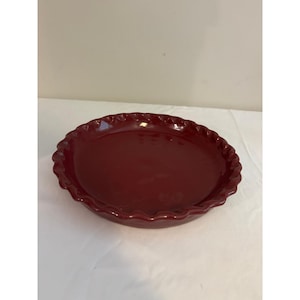 May include: A deep red ceramic pie dish with a glossy finish and a decorative scalloped edge. The round baking vessel features a smooth, reflective interior surface and a consistent dark crimson color throughout the entire exterior and rim.