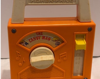 May include: An orange Fisher-Price toy radio with a cream-colored handle and central dial. The front features a graphic labeled The Candy Man, musical note symbols, and a vertical slider with numbers 55, 60, 70, 80, and 160 printed on it.