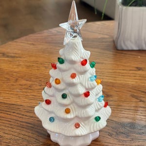 May include: A white ceramic tabletop tree with a glossy finish, featuring molded branches and small, multi-colored translucent plastic bulbs in red, green, orange, and blue. A clear, star-shaped topper sits at the peak of the tree.