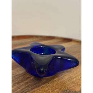 May include: A star-shaped candle holder made of deep cobalt blue glass. The object features five rounded points and a circular indentation in the center designed to hold a small candle. The glass has a smooth, polished, and reflective surface.