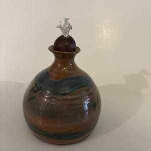 May include: A round, glazed ceramic oil lamp with a bulbous body featuring horizontal bands of brown and dark blue. The lamp has a narrow neck topped with a dark brown, spherical stopper and a white, fibrous wick protruding from the center.