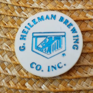 May include: A white, circular emblem with the text "G. HEILEMAN BREWING CO. INC." in blue. The emblem features a blue eagle design within a shield shape. The background is a woven straw texture.