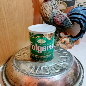 May include: A green and white Folgers Coffee can with the text "Mountain Grown Coffee" and "Naturally Decaffeinated." The can is a trial size, net weight 3.25 oz. It sits on a metal surface.
