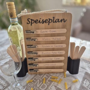 Könnte beinhalten: Ein hölzerner Speiseplan mit dem Wort "Speiseplan" oben. Er hat Schlitze für jeden Wochentag, mit Holzstäbchen für Mahlzeiten. Eine Flasche Weißwein und ein Weinglas befinden sich links. Nudeln sind auf dem Tisch verstreut.
