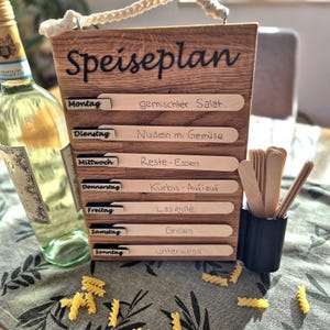 Könnte beinhalten: Ein rechteckiges hölzernes Speiseplan-Board mit der Aufschrift Speiseplan oben. Es verfügt über sieben horizontale Schlitze, die mit den Wochentagen von Montag bis Sonntag beschriftet sind und jeweils einen Holzstab mit handschriftlichen Mahlzeiten enthalten.