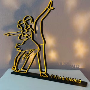 Könnte beinhalten: Goldene Silhouette eines tanzenden Paares, mit den Namen "Silvia & Harald" auf einem schwarzen Sockel.