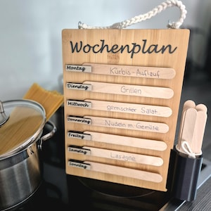 Könnte beinhalten: Ein rechteckiges Wochenplan-Board aus Bambus mit der Aufschrift Wochenplan oben. Es verfügt über sieben horizontale Schlitze für jeden Wochentag, beschriftet von Montag bis Sonntag, mit herausnehmbaren Holzstäbchen für handschriftliche Notizen.