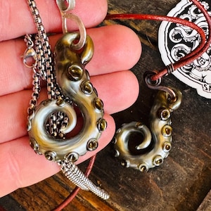 May include: Two octopus tentacle pendants. One is on a silver chain, the other on a red leather cord. The pendants are a mix of gold, silver, and brown, with small, round details. The background is a wooden surface.