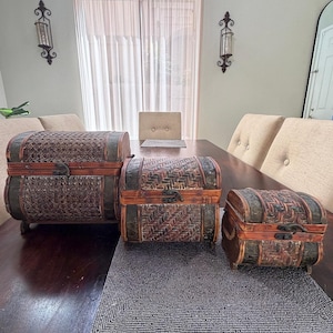 May include: A set of three graduated storage chests with arched lids, constructed from dark brown woven material and wood trim. Each chest features a metal latch on the front, decorative metal corner accents, and small feet for stability.