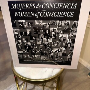 May include: A book titled "MUJERES de CONCIENCIA WOMEN of CONSCIENCE" with a black and white collage of images. The author and photographer's name, Victoria G. Alvarado, is printed at the bottom. The book is displayed on a gold and white table.