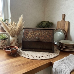 May include: A dark brown wooden bread box with a slanted front panel featuring the word Bread in a light-colored script font. The box has a single round knob on the front panel and a rectangular base, showcasing a visible wood grain texture.