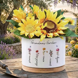 Op de afbeelding: Een witte keramische bloempot met de tekst "Grandma's Garden" en namen onder kleurrijke bloemenillustraties. De pot bevat heldergele zonnebloemen. De basis is van hout.