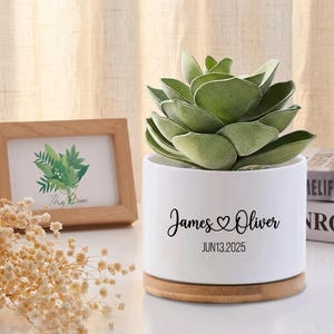 May include: A white ceramic planter with a wooden base, holding a green succulent plant. The planter is inscribed with "James ♡ Oliver" and the date "JUN13.2025". A small framed picture and dried flowers are in the background.