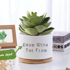 Puede incluir: Una maceta de cerámica blanca con base de madera, con la inscripción "Grow With The Flow". Contiene una planta suculenta verde. Una impresión botánica enmarcada y libros apilados se encuentran en el fondo.