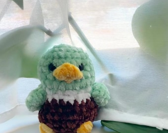 May include: A handmade crochet duckling plushie. The duckling has a mint green head and wings, a white chest, and a dark brown body. It features a yellow beak and feet, and black eyes. The plushie is a cute, soft toy.