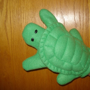 May include: Green felt hand puppet shaped like a turtle with black eyes. The puppet has a textured shell.