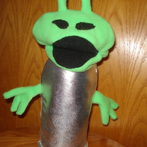 May include: A green alien puppet with a silver body and a large black mouth. The puppet has two antennas on its head and is made of soft fabric.