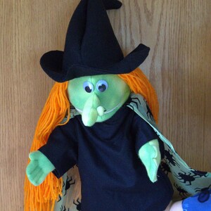 May include: A handmade witch puppet with a black hat, green face, and orange yarn hair. The witch wears a black shirt and a cape with a spider pattern. The puppet has blue eyes and a long nose.