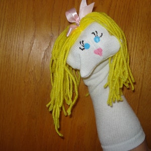May include: A white sock puppet with yellow yarn hair, blue eyes, and a pink nose. The puppet is wearing a pink bow.