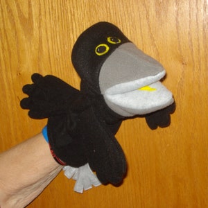 May include: A black bird hand puppet featuring a large grey beak, yellow eyes, and a small yellow tongue. The puppet has black wings and a white tail, constructed from soft fabric. The design includes a movable mouth and detailed facial features.