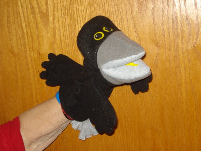 May include: A black bird hand puppet featuring a large grey beak, yellow eyes, and a small yellow tongue. The puppet has black wings and a white tail, constructed from soft fabric. The design includes a movable mouth and detailed facial features.