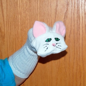 May include: White sock puppet with pink ears and green eyes, shaped like a cat. The puppet has a white face with a pink nose and black whiskers.