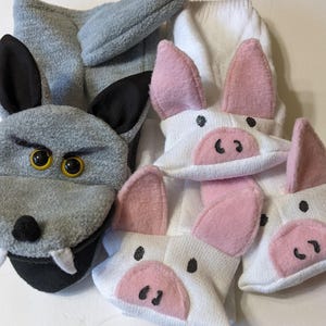 May include: A grey fleece wolf puppet with black ears, yellow eyes, and white fangs, positioned next to three white knit socks featuring pink felt pig ears and snouts with black stitched details, creating a set of animal-themed character accessories.
