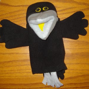 May include: A black bird hand puppet made of soft fabric with outstretched wings. The puppet features a grey beak, a bright yellow inner beak, and two yellow eyes with black pupils. At the bottom, there are two light grey fabric feet.