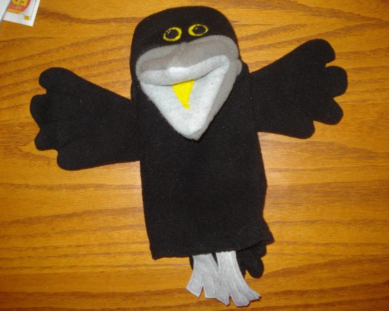 May include: A black bird hand puppet made of soft fabric with outstretched wings. The puppet features a grey beak, a bright yellow inner beak, and two yellow eyes with black pupils. At the bottom, there are two light grey fabric feet.