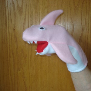 May include: Pink felt hand puppet shaped like a shark with a white felt mouth and red felt teeth. The puppet is on a person's hand.