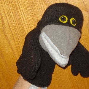 May include: A black crow hand puppet made of soft fabric. The bird features a large grey beak, a light grey lower jaw, and two round yellow eyes with black pupils. The puppet has two small black wings extending from the sides of its body.